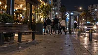 ספסל ברחוב לילי ליד מסעדה, עם אבנים על המדרכה וקבוצת צעירים מטושטשת ברקע