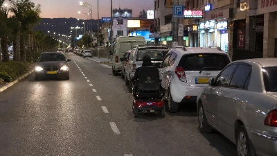כביש עירוני באילת בשעת ערב עם קלנועית סמוך לשורת כלי רכב חונים ונתיב שמאלי פנוי